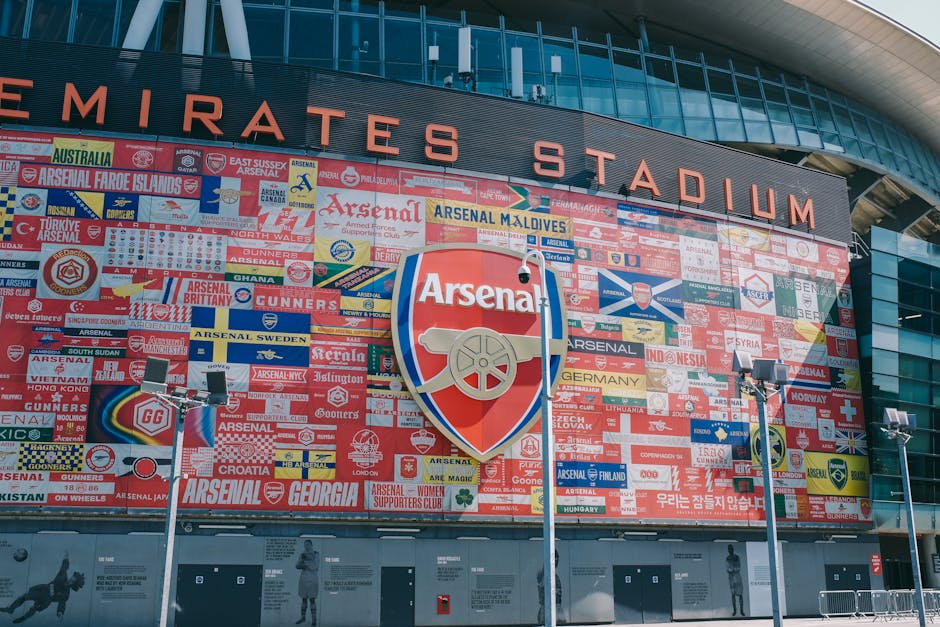 Exterior view of Emirates Stadium with a large wall display featuring numerous colorful Arsenal football club badges and signs, including the prominent Arsenal shield logo in the center, under a modern curved glass facade. The building's surface is clean, showcasing reflective glass panels and a neatly maintained mosaic of team insignias, with bright outdoor lighting illuminating the scene. The image highlights the stadium's architectural design and the decorative sports-related artwork outside, providing a vibrant and detailed perspective on the exterior environment.