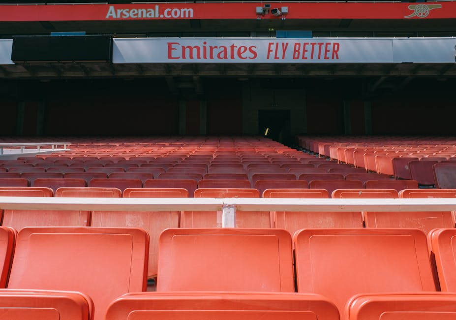 Inside the Emirates Stadium with rows of red plastic seating extending toward a dark tunnel in the background. The seats appear clean and well-maintained, with no visible dust or dirt. Above the seating area, a large sign displays the Emirates logo along with the slogan 'Fly Better,' set against a red and white background. The lighting highlights the smooth, shiny surface of the seats, indicating recent surface cleaning or sanitisation. The structure features metal supports and a section of the stadium’s roof, contributing to the clean and professional appearance associated with high-quality domestic and commercial cleaning standards, as represented by Carpet Cleaners N5 on the page titled 'Emirates Stadium upholstery cleaning and stain tips Highbury, N5,' emphasizing surface cleaning and maintenance techniques.