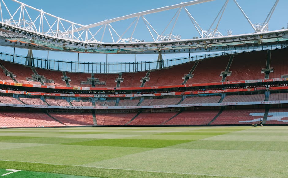 Exterior view of Emirates Stadium with a large wall display featuring numerous colorful Arsenal football club badges and signs, including the prominent Arsenal shield logo in the center, under a modern curved glass facade. The building's surface is clean, showcasing reflective glass panels and a neatly maintained mosaic of team insignias, with bright outdoor lighting illuminating the scene. The image highlights the stadium's architectural design and the decorative sports-related artwork outside, providing a vibrant and detailed perspective on the exterior environment.